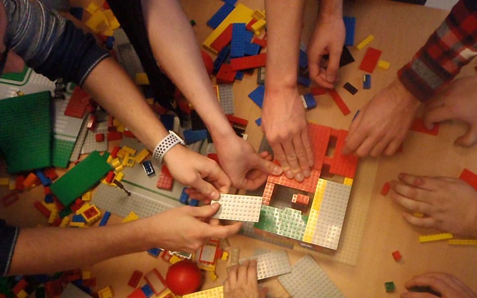 Foto: Auf einem Tisch sind viele Hände zu sehen und bauen etwas mit Legoteilen zusammen.