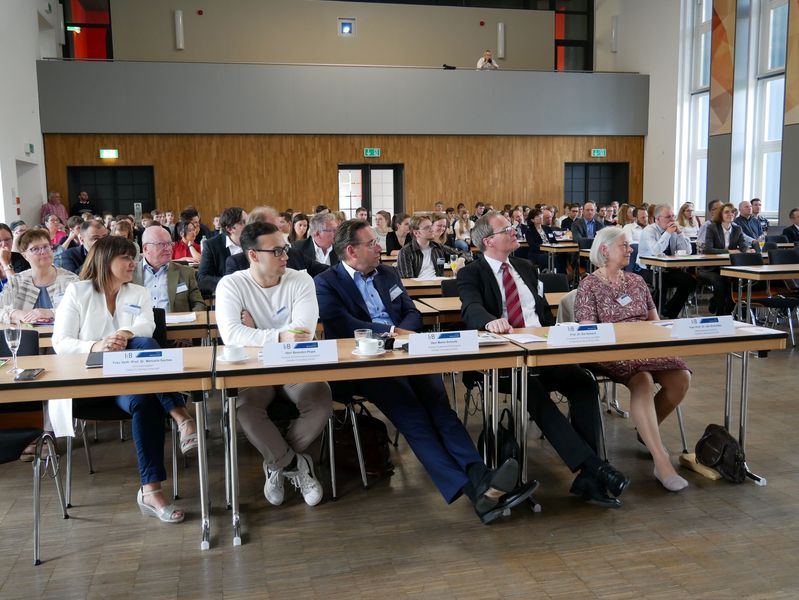 Foto: In einer großen Aula sitzen viele Menschen an Tischen.