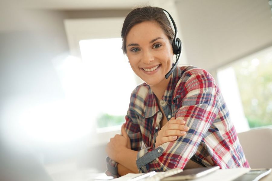 Foto: Eine Frau sitzt mit Headset am Schreibtisch und lächelt in die Kamera.