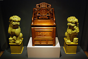 Foto: Ein verzierter Stuhl aus Holz auf einem Sockel. Links und rechts daneben jeweils ein chinesischer Wächterlöwe auf einem Sockel.