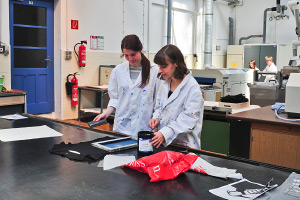 Foto: Zwei Studierende bearbeiten an einem Arbeitstisch einen Stoff mit Farbe.