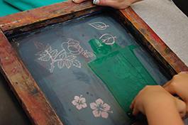 Foto: Zwei Hände tätigen einen Kunstdruck in einem Holzrahmen auf ein Textil.