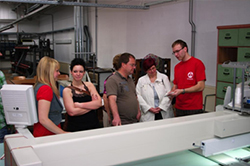 Foto: 6 Personen stehen vor einer Maschine. Student Alexander Wille (rechts) erklärt interessierten Besuchern die Möglichkeiten einer modernen Mehrkopfstickmaschine. 