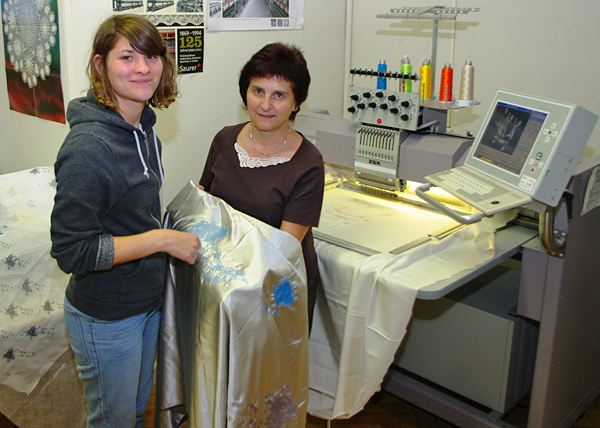 Foto: Studentin Stefanie Mittmann mit Frau Gisela Mertinat an der Stickmaschine. Sie zeigen einen langen Stoff mit Muster in die Kamera.