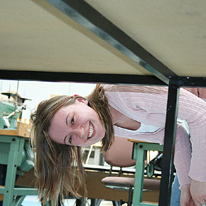 Foto: Eine Studierende schaut gebeugt unter einem Tisch hindurch und lächelt in die Kamera.