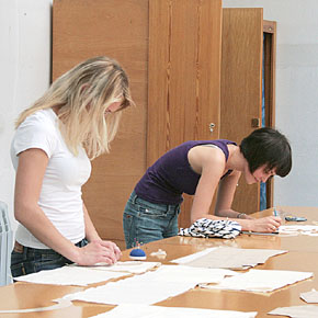 Foto: Zwei Studierende tätigen an einem Tisch stehend Zuschnitte.