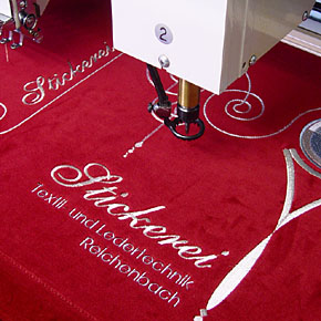 Foto: Eine Stickmaschine stickt Schriftzüge und ein Muster in roten Stoff.