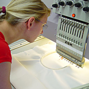 Foto: Eine Studierende betrachtet mehrfach parallel verlaufenden Nadeln einer Stickmaschine bei ihrer Arbeit.
