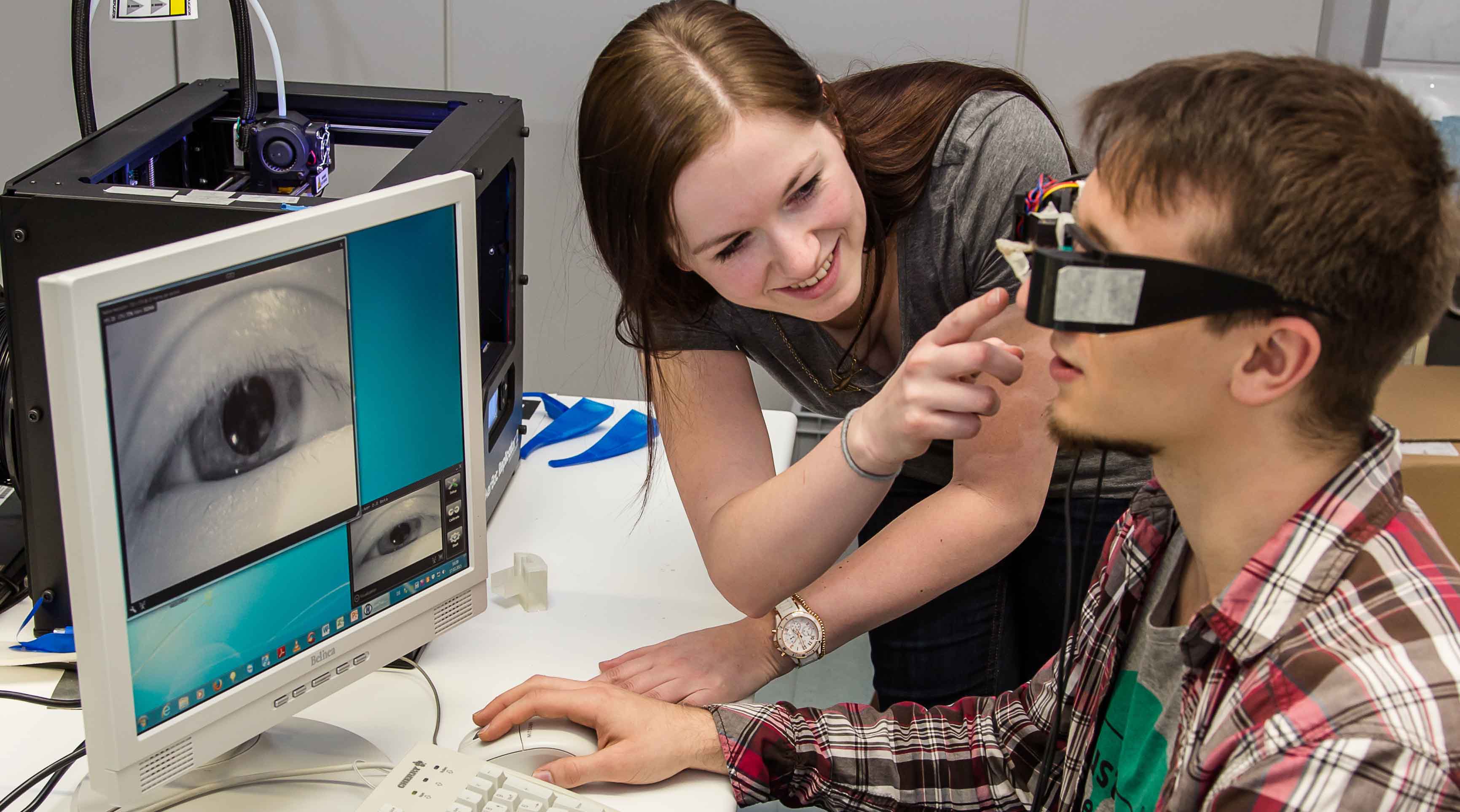 Foto: Ein Student trägt eine Brille mit Kamera. Auf einem Bildschirm vor ihm ist sein Auge zu erkennen. Eine Studierende steht daneben und assistiert.