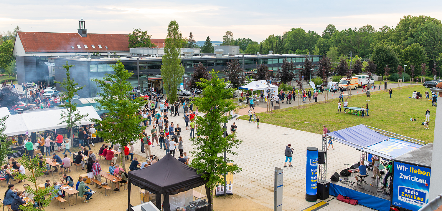 Blick über den Campus Scheffelstraße, ein schöner Sommerabend, im Vordergrund Bühne und Grillstand, im Hintergrund Laborgebäude, viele Familien spazieren über den Campus. 