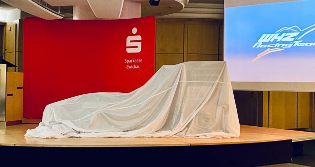 Blick auf die Bühne mit rotem Hintergrund und Logo der Sparkasse. Auf der Bühne steht weiß verhüllt der neue Rennwagen.