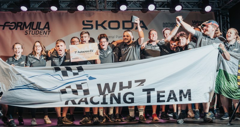 Die Mitglieder des WHZ Racing Teams jubelnd nach einem Sieg. Sie halten einen Banner mit dem Logo des Racing Teams in der Hand