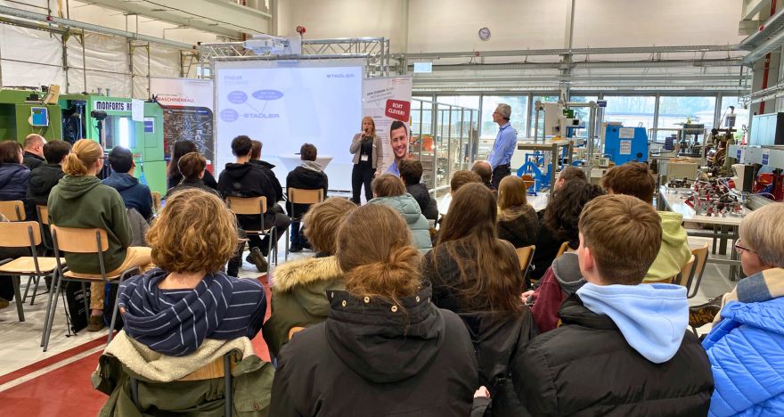 Maschinenhalle mit Blick auf Leinwand und Redner, im Vordergrund sind die Zuschauer zu sehen