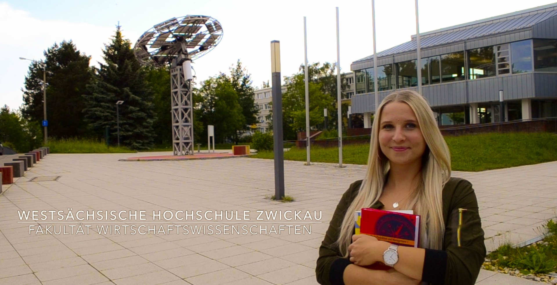 Videovorschaubild. Link zum Video. Eine Studierende steht auf dem Vorplatz zum Campus Scheffelberg. Im Hintergrund ist ein Solarkollektorturm und das Gebäude Mensa Scheffelberg zu sehen.