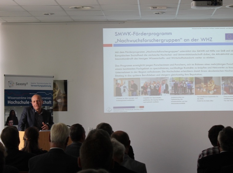 Foto: Teilnehmer sitzen zu einem Vortrag im Seminarraum zum IfP Symposium. Ein Redner präsentiert eine Powerpoint.