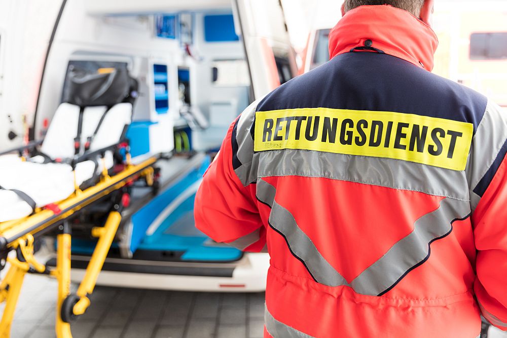 Foto: Im Vordergrund steht ein Mann mit dem Rücken zur Kamera. Auf seiner Jacke steht groß Rettungsdienst. Im Hintergrund ist - verschwommen zu sehen - ein offener Krankentransportwagen.