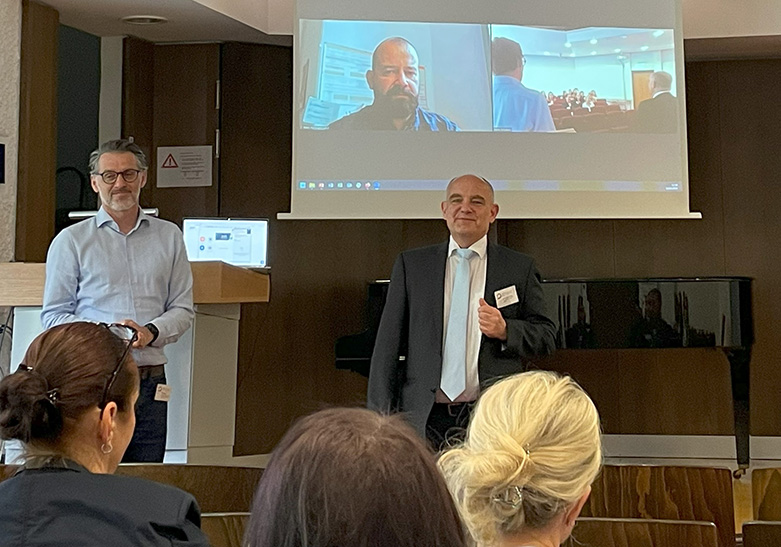 zwei männliche Referenten live und ein Referenz online per Videoschalte vor Publikum