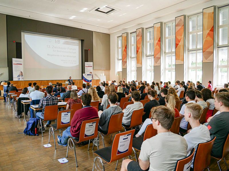 Menschen in einer Aula platziert auf roten Stühlen mit Blick auf die Bühne