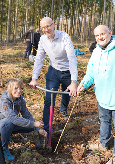 Drei Personen stehen in einem Wald und blicken in die Kamera, eine Person sticht mit einem Spaten in die Erde. 