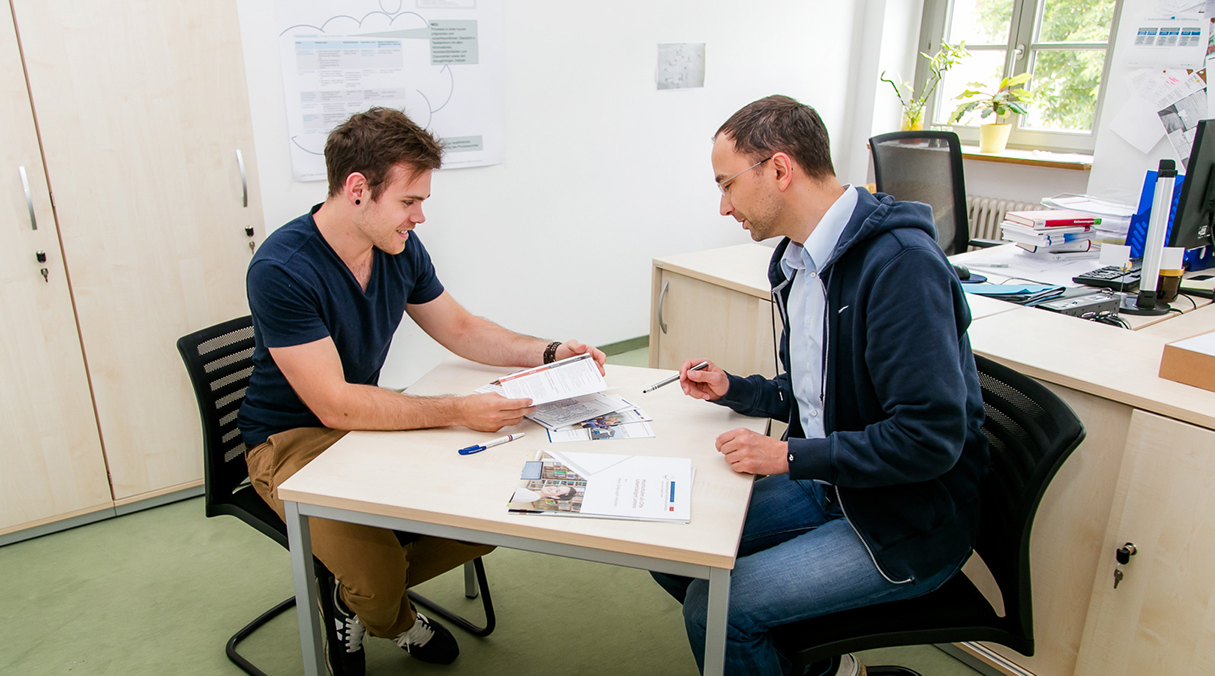 An einem Tisch sitzend berät ein Studienberater einen Studieninteressenten.