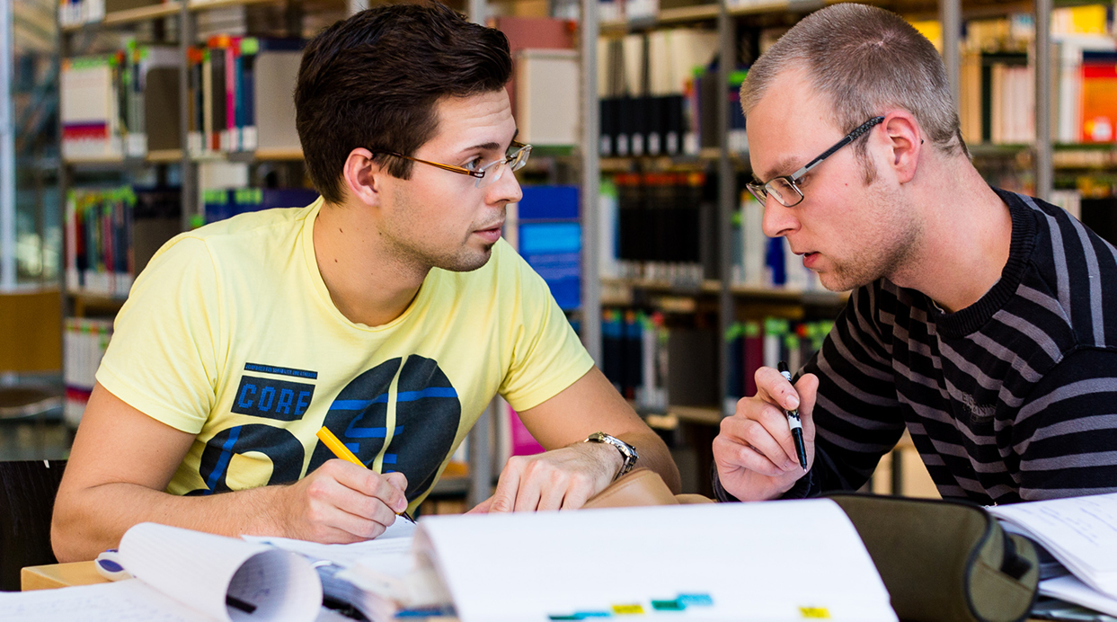 Zwei Studierende sitzen mit ihren Arbeitsmaterialien in der Bibliothek an einem Tisch und diskutieren miteinander.