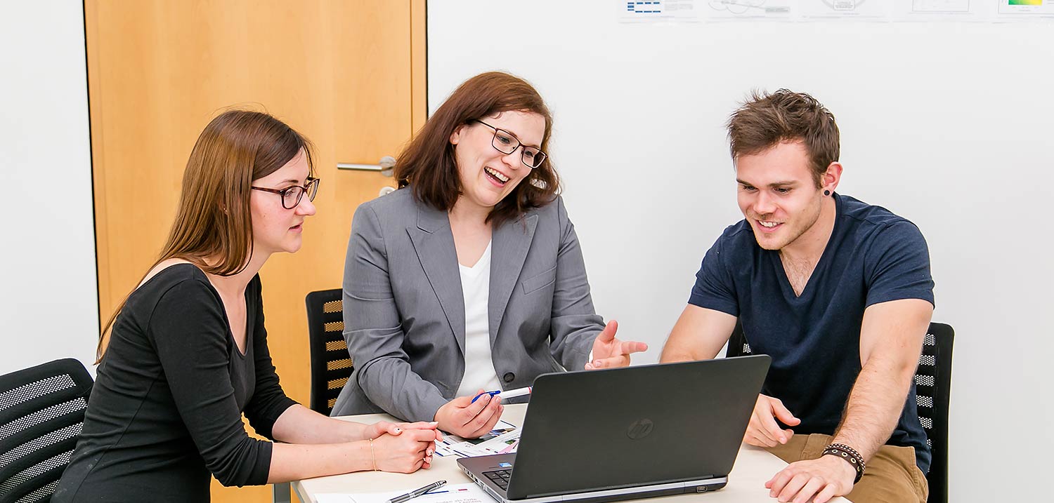 Foto: Eine Frau berät am Tisch mit einem Laptop zwei Studieninteressenten.