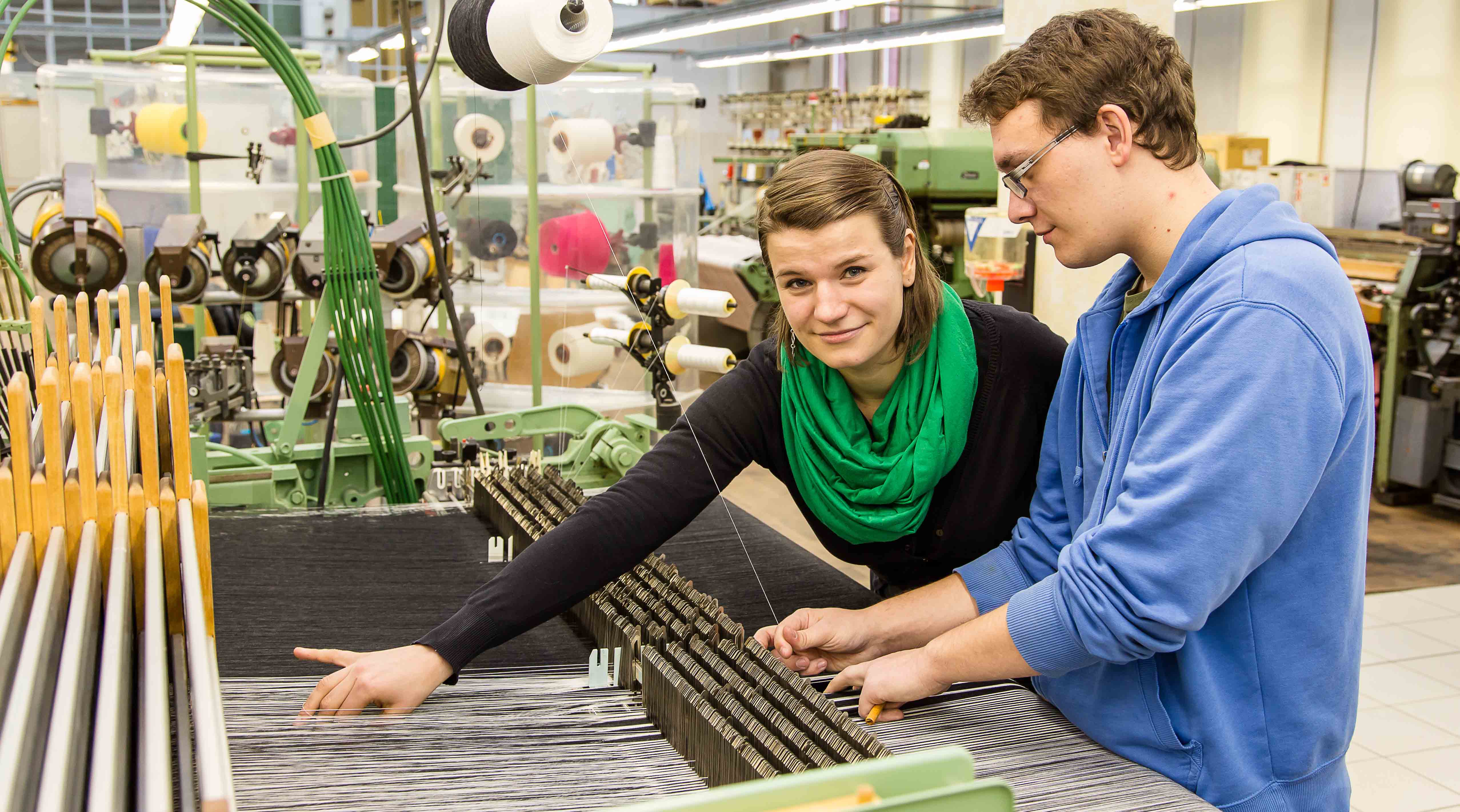 Foto: Zwei Studierende arbeiten an einer Maschine worin Fäden gespannt sind. Textile Strukturen und Technologien (Bachelor).