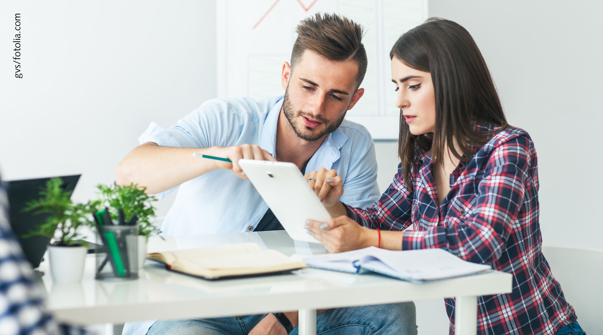 Foto: Zwei Studierende sitzen an einem Tisch und besprechen etwas. Dabei schauen Sie gemeinsam auf den Bildschirm eines Tablet.