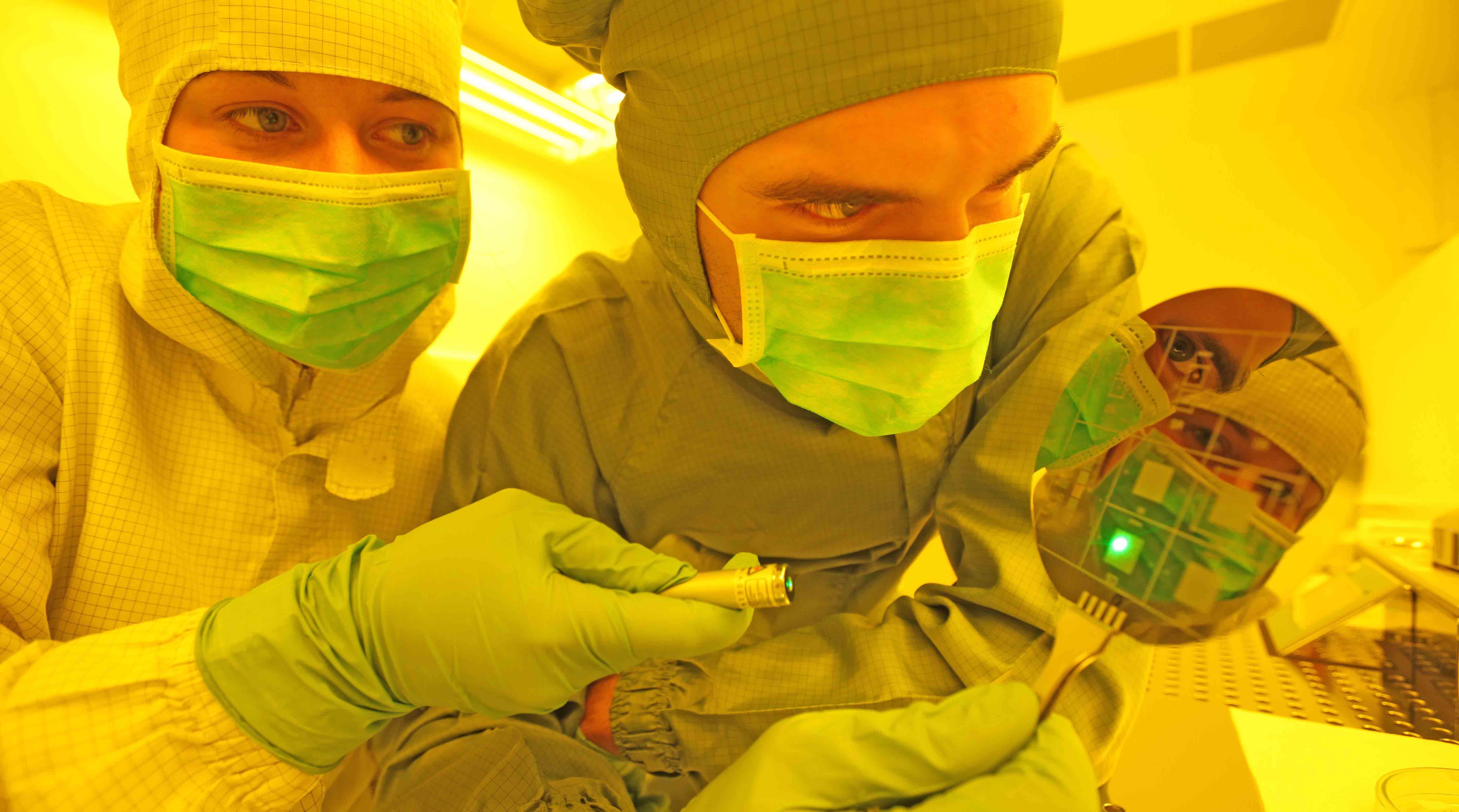 Foto: Zwei Personen in Schutzkleidung mit Kopfbedeckung und medizinischen Mundschutz in einem Labor. Eine Person hält einen ovalen Gegenstand in der Hand, dessen Oberfläche wie ein Spiegel wirkt und die Gesichter darin zu erkennen sind.