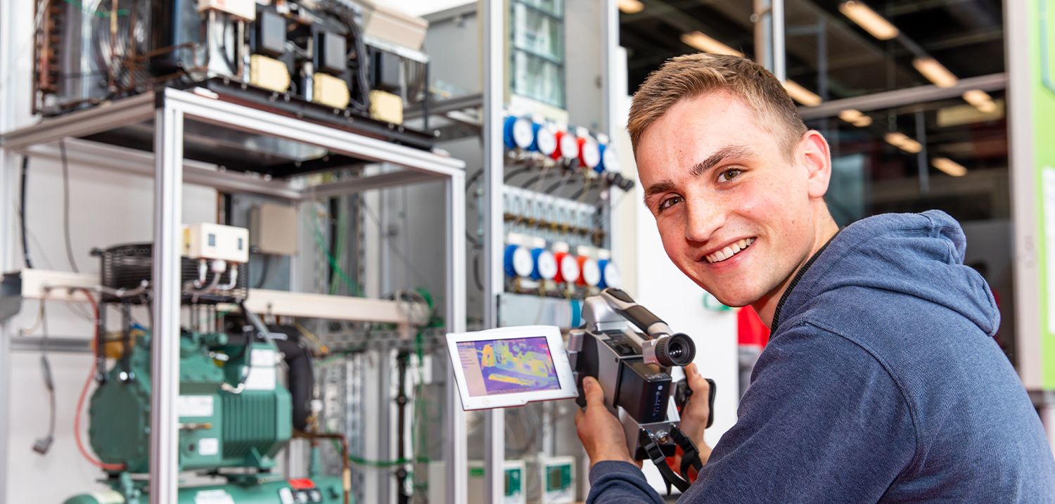 Student blickt in die Kamera, hält eine Wärmebildkamera in der Hand, im Hintergrund eine Laborhalle mit Apparaturen