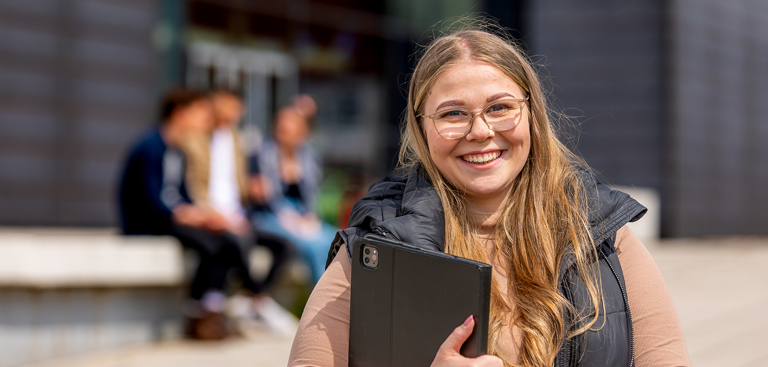 Eine Studierende mit Tablet steht auf dem Campus und blickt in die Kamera. 