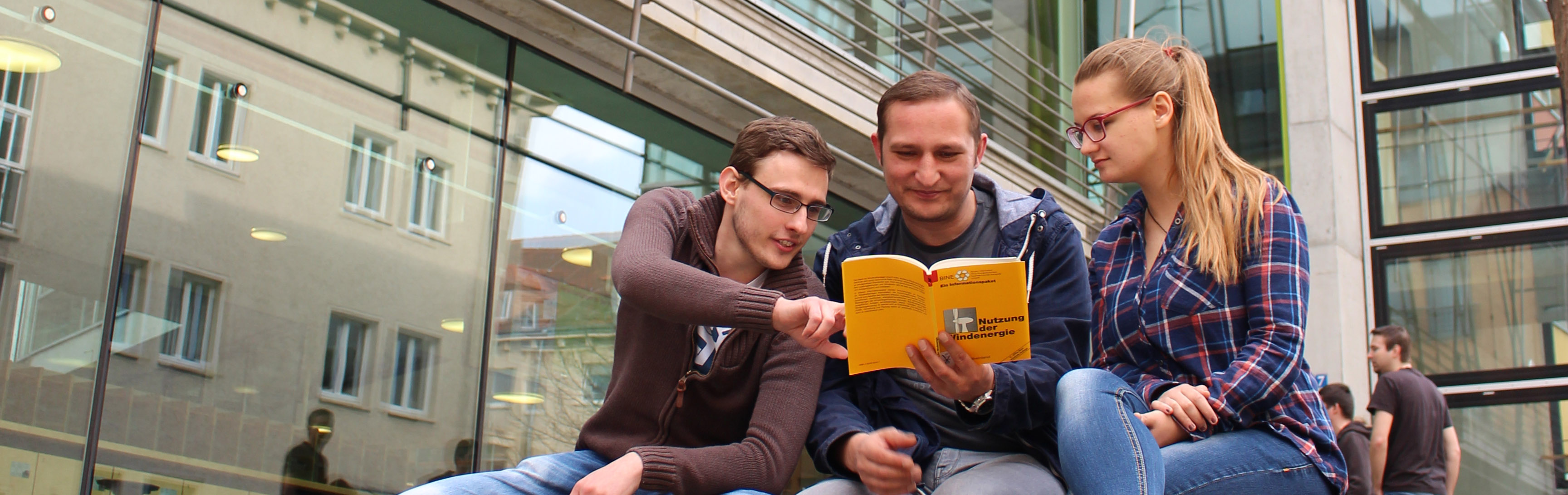 Foto: Drei Studierende sitzen vor der Hauptbibliothek und schauen gemeinsam in ein Buch zur Nutzung der Windenergie.